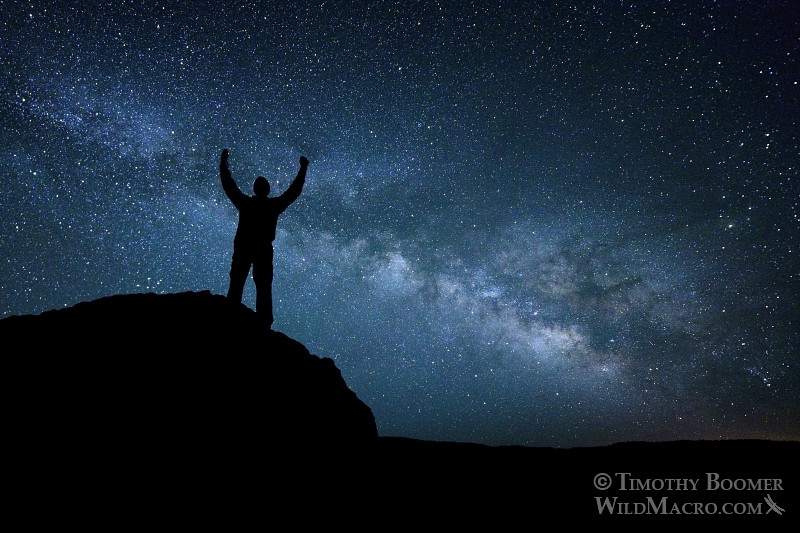 A night hiker stands triumphantly atop a massive boulder under the splendor of the Milky Way Galaxy.  Eldorado National Forest, El Dorado County, CA.  Stock Photo ID=SCE0106